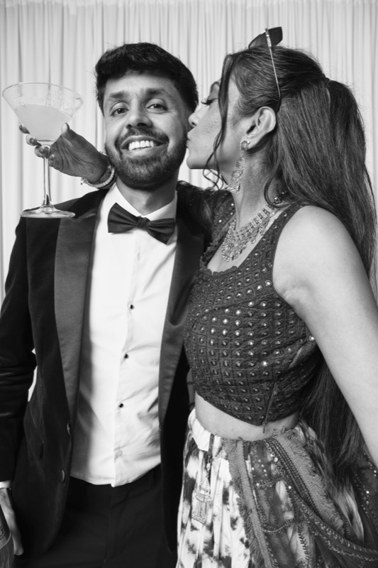 A woman kisses a smiling man in a bow tie, a martini glass on his shoulder
